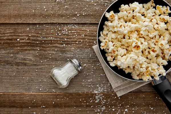 Top view of delicious popcorn in frying pan near scattered salt on wooden background — Stock Photo