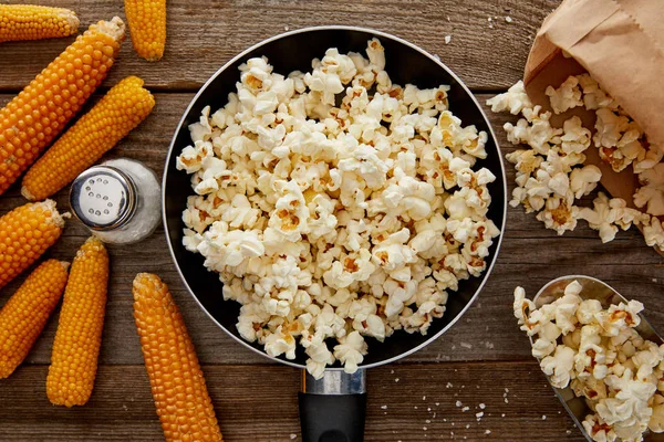 Top view of salty popcorn in frying pan near corn on wooden background — Stock Photo