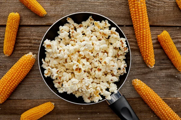 Vista dall'alto di gustosi popcorn in padella vicino a mais sparso su sfondo di legno — Foto stock