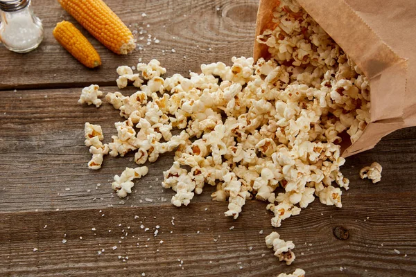 Salty delicious popcorn scattered from paper bag near corn on wooden background — Stock Photo