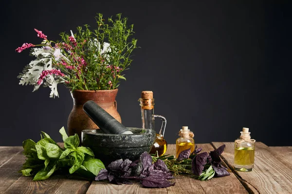 Argamassa cinzenta perto de vaso de argila com ervas e pequenas garrafas em mesa de madeira isolada em preto — Fotografia de Stock