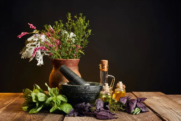 Mortero gris cerca de jarrón de arcilla con flores silvestres frescas y hierbas sobre mesa de madera aislada en negro - foto de stock