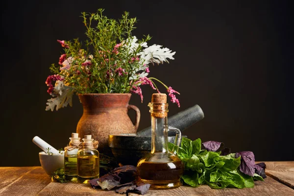 Vaso di argilla con erbe fresche e fiori vicino malta e pestello e bottiglie su tavolo di legno isolato su nero — Foto stock