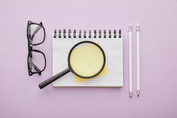 Top view of magnifier on notebook near glasses and pencils isolated on purple — Stock Photo