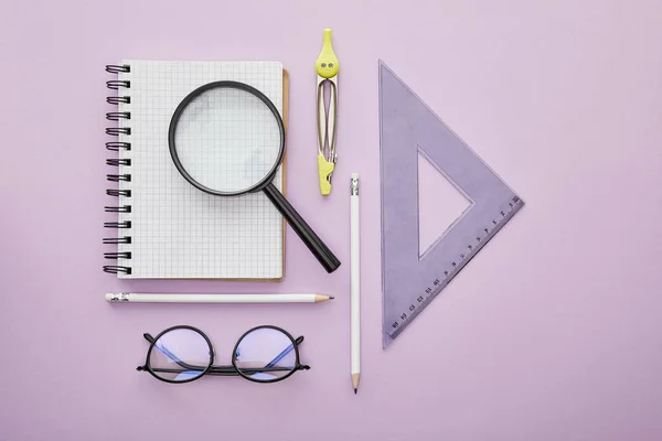 Top view of ruler triangle near magnifier on notebook near pencils, drawing compass and glasses isolated on purple — Stock Photo