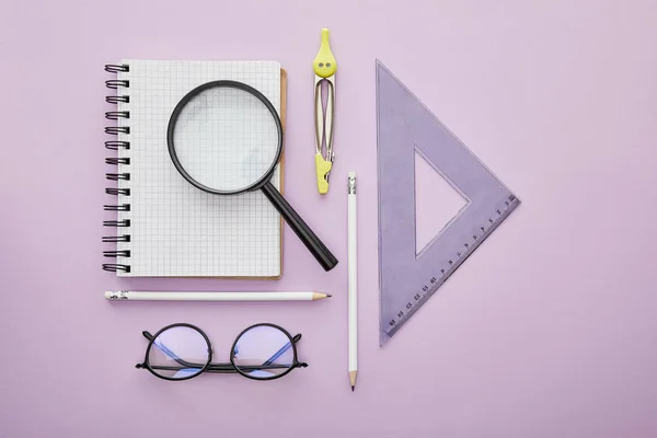 Vue du haut du triangle de la règle près de la loupe sur ordinateur portable près des crayons, dessin boussole et verres isolés sur violet — Photo de stock