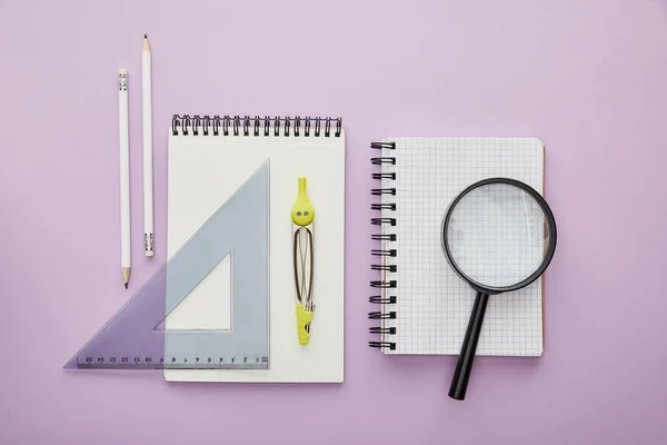 Top view of ruler triangle and drawing compass on notebook near pencils and magnifier isolated on purple — Stock Photo