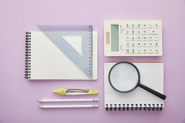 Top view of magnifier and ruler triangle on notebooks near calculator and stationery isolated on purple — Stock Photo
