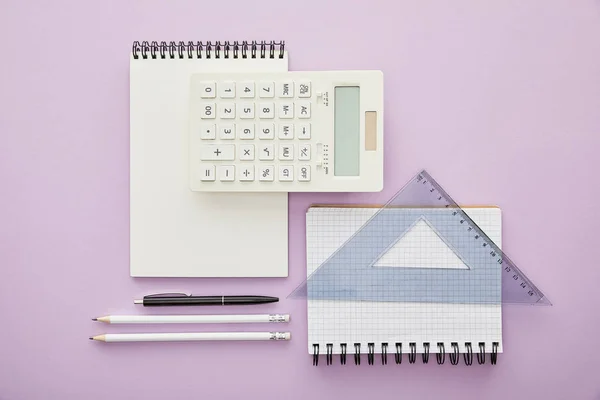 Vista superior do triângulo da régua no caderno perto da calculadora e artigos de papelaria isolados no roxo — Fotografia de Stock