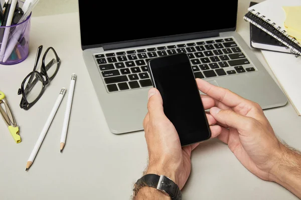 Обрізаний вид людини, що тримає смартфон з порожнім екраном біля ноутбука та канцелярських товарів на білому — стокове фото