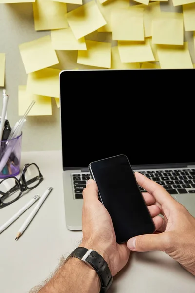 Vista cortada do homem segurando smartphone com tela em branco perto de laptop, óculos e artigos de papelaria em cinza e branco — Fotografia de Stock