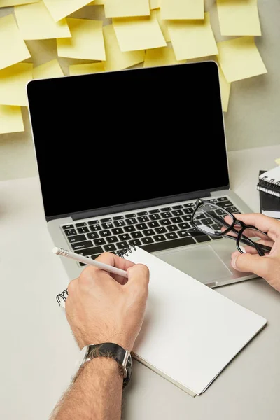 Cropped view of man holding glasses while writing on notebook near laptop with blank screen on grey and white — Stock Photo