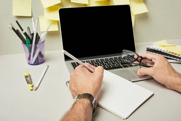 Cropped view of man holding glasses while writing on notebook near laptop with blank screen and stationery on grey and white — Stock Photo