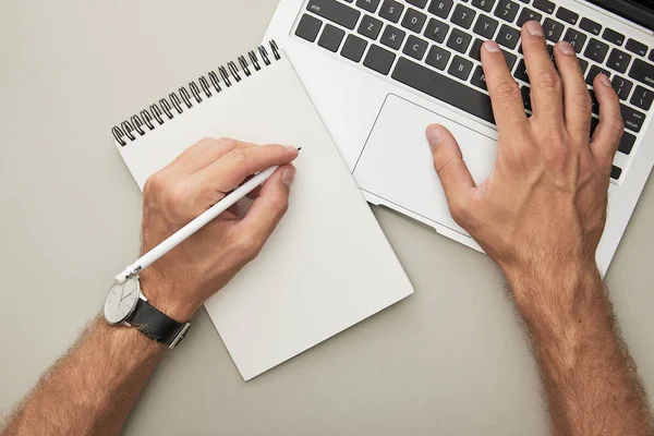 Vista recortada del hombre usando el ordenador portátil mientras sostiene el lápiz cerca del cuaderno aislado en gris - foto de stock