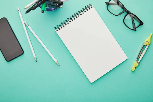 Top view of smartphone with blank screen near glasses, stationery and notebook isolated on turquoise — Stock Photo