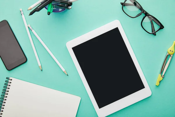 Top view of gadgets with blank screens near glasses, stationery and notebook isolated on turquoise — Stock Photo