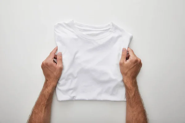 Vista recortada del hombre sosteniendo camiseta plegada en blanco sobre fondo blanco - foto de stock
