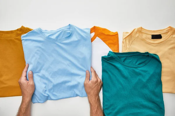 Cropped view of man holding blue t-short near beige, orange, turquoise and ochre ones on white background — Stock Photo