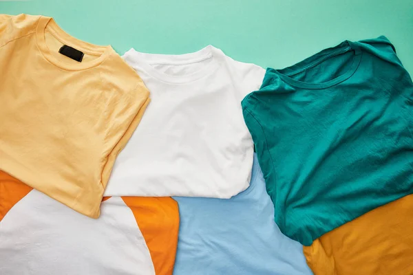Вид сверху на сложенные оранжевые, бежевые, белые, голубые и охристые футболки на бирюзовом фоне — стоковое фото