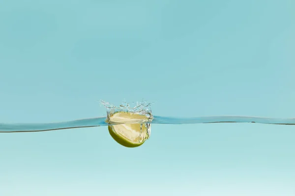 Medio de cal madura cayendo en el agua con salpicadura sobre fondo azul - foto de stock