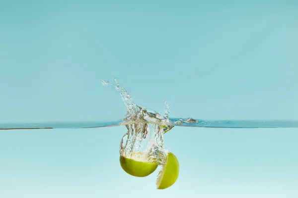 Reife Kalkhälften fallen in Wasser mit Spritzer auf blauem Hintergrund — Stockfoto