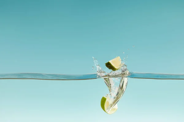 Mitades de cal maduras que caen profundamente en el agua con salpicaduras sobre fondo azul - foto de stock