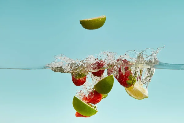 Reife Limettenstücke und Erdbeeren fallen tief ins Wasser mit Spritzer auf blauem Hintergrund — Stockfoto