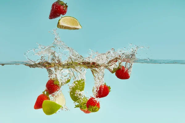 Pedaços de limão e morangos caindo profundamente na água com respingo no fundo azul — Fotografia de Stock