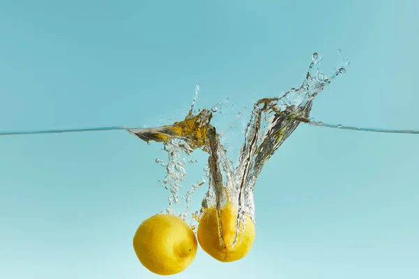 Цілі стиглі лимони глибоко падають у воду з бризкою на синьому фоні — стокове фото