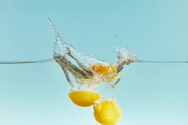 Gelbe Zitronen fallen tief ins Wasser mit Spritzer auf blauem Hintergrund — Stockfoto