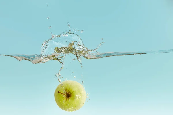 Зелене яблуко глибоко падає у воду з бризкою на синьому фоні — стокове фото