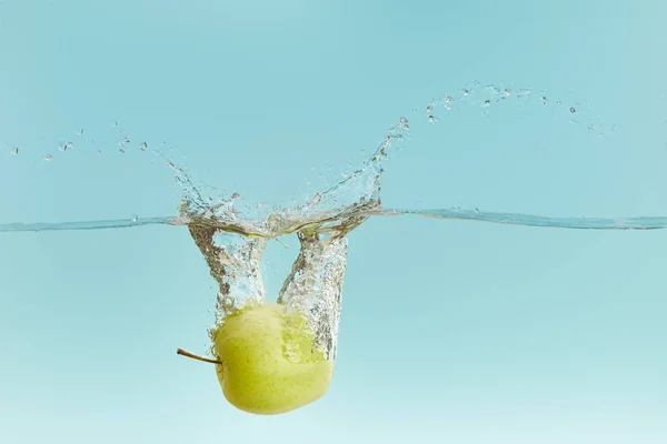 Стигле зелене яблуко глибоко падає у воду з бризкою на синьому фоні — стокове фото