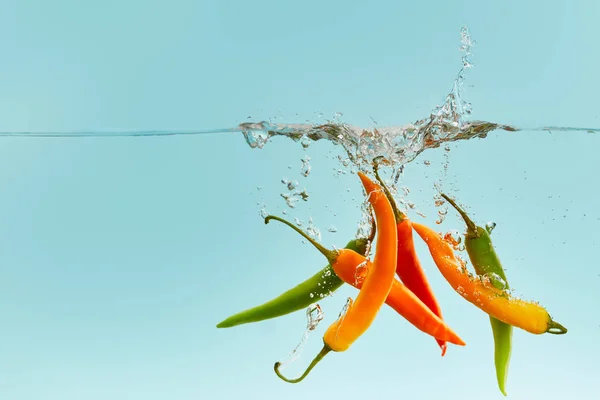 Bunte Chilischoten fallen tief ins Wasser mit Spritzer auf blauem Hintergrund — Stockfoto