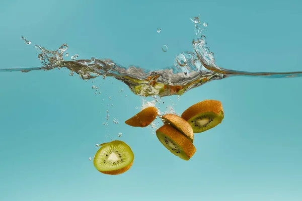 Rodajas de kiwi que caen profundamente en el agua con salpicaduras en el fondo azul - foto de stock