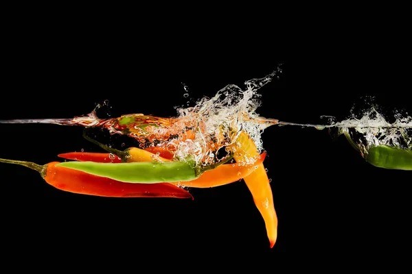 Pimentas multicoloridas que caem profundamente na água com respingo isolado em preto — Fotografia de Stock