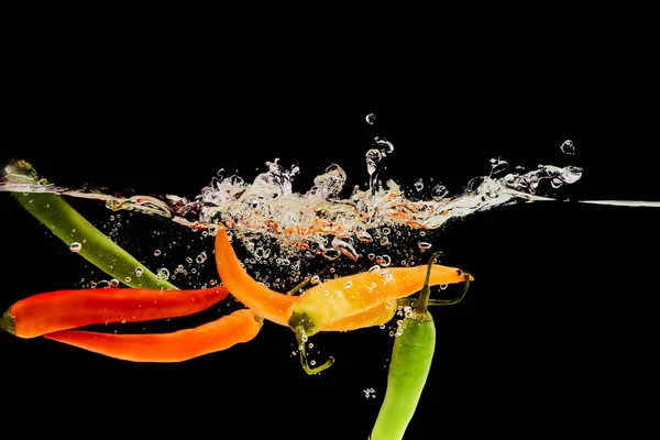 Bunte Chilischoten fallen tief ins Wasser mit Spritzer isoliert auf schwarz — Stockfoto