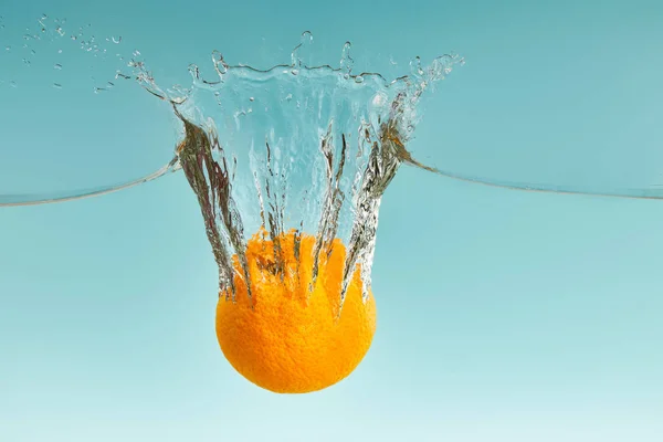 Leckere frische Orange fällt in Wasser mit Spritzer auf blauem Hintergrund — Stockfoto