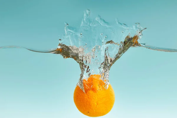 Стиглий свіжий апельсин падає у воду з бризкою на синьому фоні — стокове фото