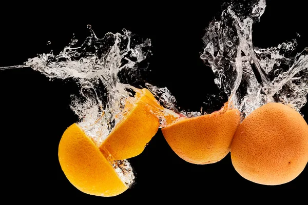 Toronja madura y mitades de naranja que caen en el agua con salpicaduras aisladas en negro - foto de stock