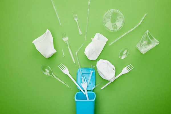 Vista dall'alto di bicchieri di plastica usa e getta, forchette e cucchiai vicino al cestino blu su sfondo verde — Foto stock