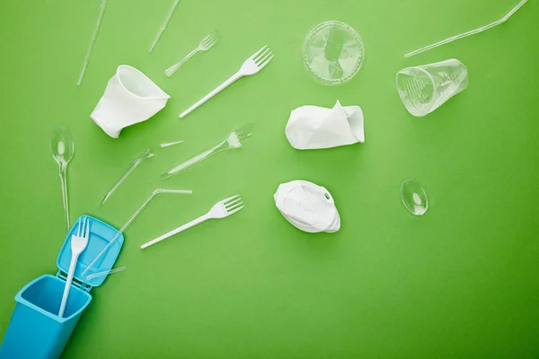 Vista superior de copas de plástico arrugado, tenedores y cucharas cerca de la papelera de reciclaje azul sobre fondo verde - foto de stock