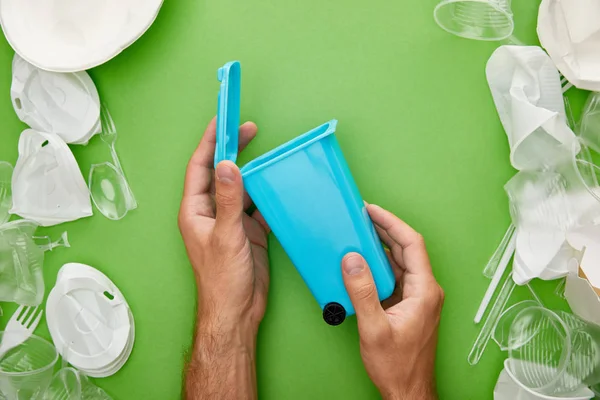 Vista recortada del hombre sosteniendo la papelera de reciclaje azul cerca de copas de plástico arrugado sobre fondo azul - foto de stock