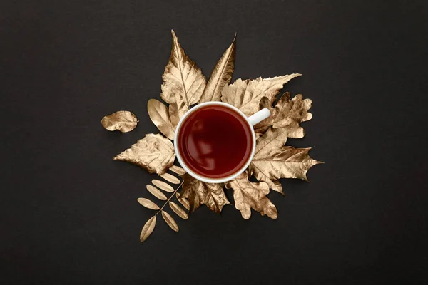 Vista superior do chá na caneca perto de folhagem dourada no fundo preto — Fotografia de Stock