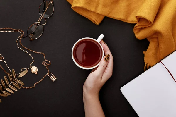 Vue partielle de la main féminine près du thé dans une tasse et accessoires automnaux sur fond noir — Photo de stock