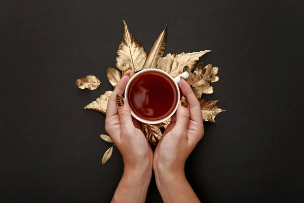 Частковий вигляд жінки, що тримає чай в кухольці біля золотого листя на чорному тлі — стокове фото