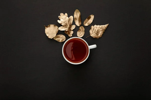Vista superior del té caliente en taza cerca del follaje dorado sobre fondo negro - foto de stock