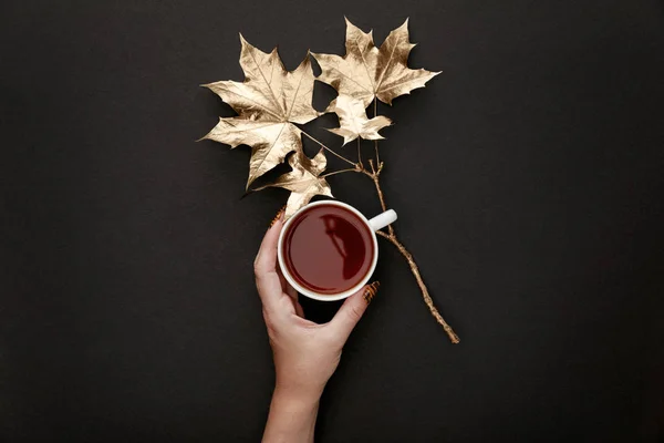 Vista ritagliata della donna che tiene il tè in tazza vicino foglie d'acero dorate su sfondo nero — Foto stock