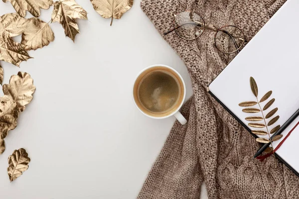 Draufsicht auf gestrickten braunen Pullover, Brille, Kaffee, leeres Notizbuch und goldenes Laub auf weißem Hintergrund — Stockfoto