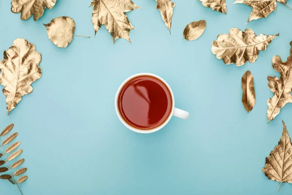 Draufsicht auf warmen Tee im Becher nahe goldenem Laub auf blauem Hintergrund — Stockfoto