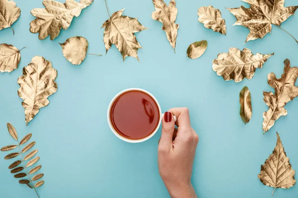 Vista ritagliata della donna che tiene il tè in tazza vicino al fogliame dorato su sfondo blu — Foto stock